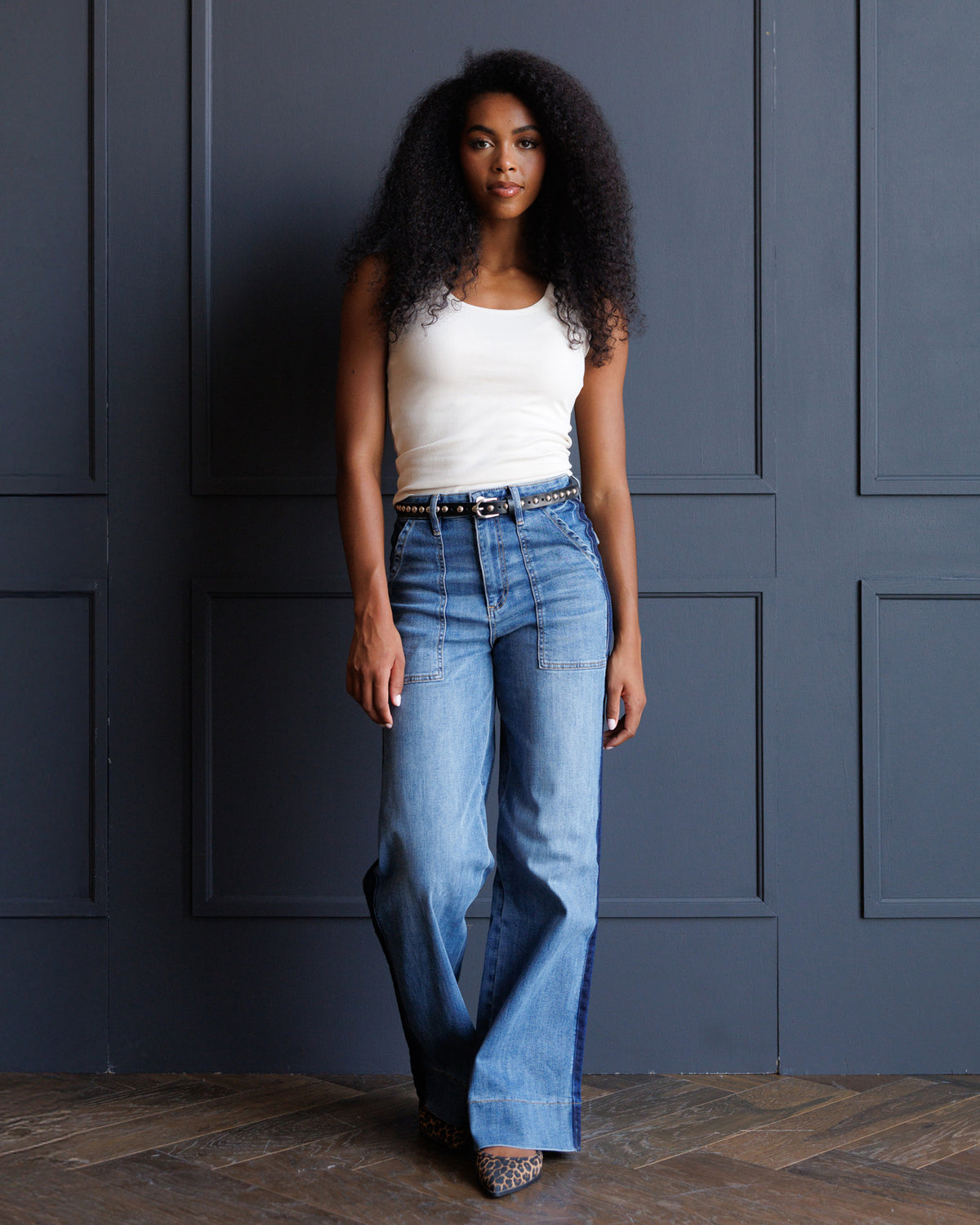 A woman with curly black hair stands against a dark blue paneled wall wearing a white sleeveless top and light blue wide-leg jeans with a black belt.