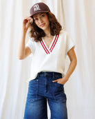 Woman wearing a white shirt with red accents, blue jeans, and a brown cap with 'PAS MAL' text against a white curtain background.