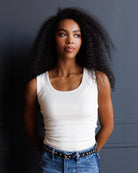 A woman with voluminous curly black hair wearing a fitted white scoop-neck tank top and blue jeans with a black studded belt, standing against a dark gray paneled wall.
