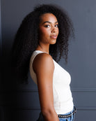 A woman with voluminous, curly black hair is shown in profile against a dark gray paneled wall. She is wearing a white sleeveless top and blue jeans with a studded belt.