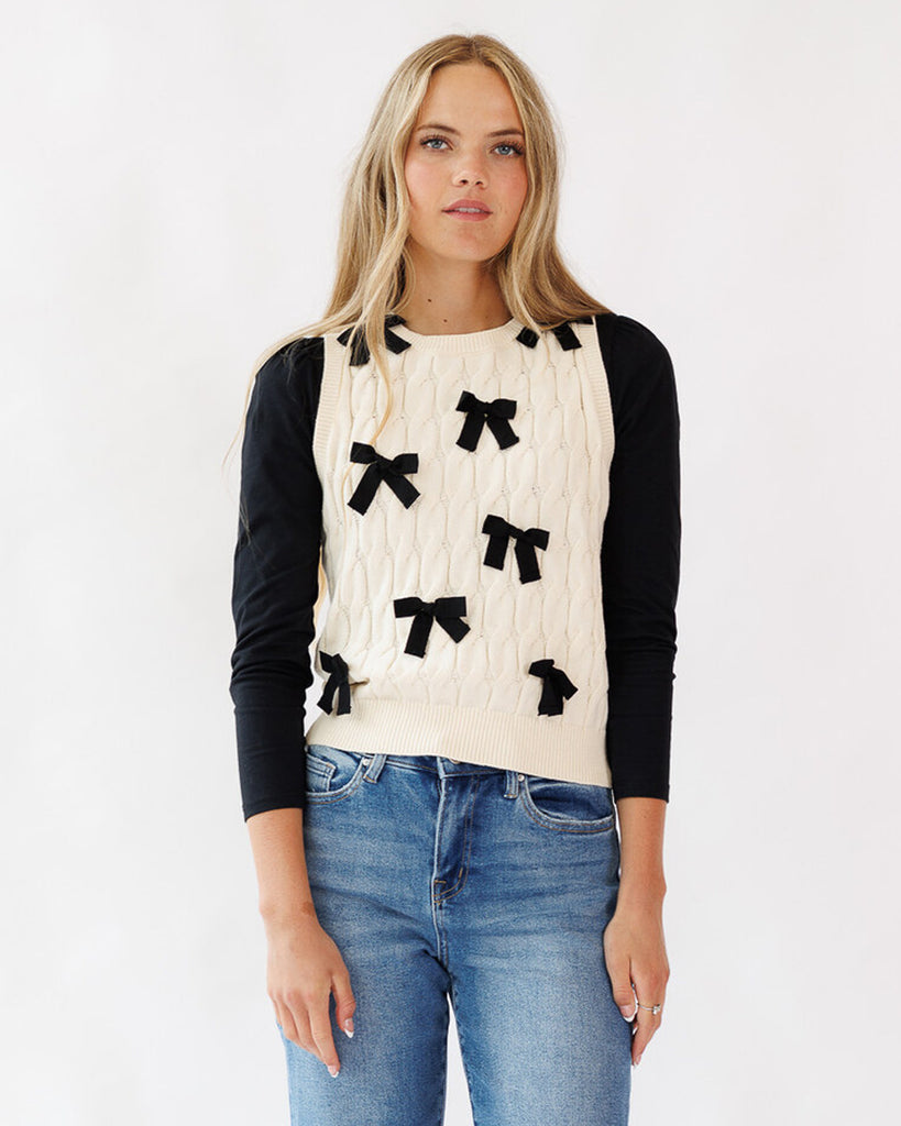 Woman wearing a cream sweater vest with black bow details and blue jeans on a white background