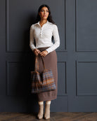 A woman stands against a dark paneled wall wearing a white and gray striped long-sleeve polo top, a brown knit midi skirt, beige socks, and beige ankle boots, while holding a brown and blue plaid tote bag.