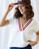 Woman wearing a white sweater with red trim and a brown cap, standing against a white background