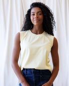 Woman wearing a light yellow sleeveless top and blue jeans against a white curtain background