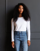 A woman with long, curly black hair stands against a dark gray paneled wall, wearing a fitted white long-sleeve crewneck top and high-waisted blue jeans with unique patch pockets.