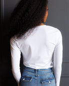 A woman with long, curly black hair is shown from behind wearing a fitted white long-sleeve t-shirt and blue jeans against a dark gray paneled wall.