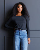 A woman with long, curly black hair stands against a dark gray paneled wall, wearing a fitted black long-sleeve crewneck top and light blue high-waisted jeans.
