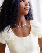 Woman wearing a white dress with puffed sleeves and a necklace, standing against a neutral background.
