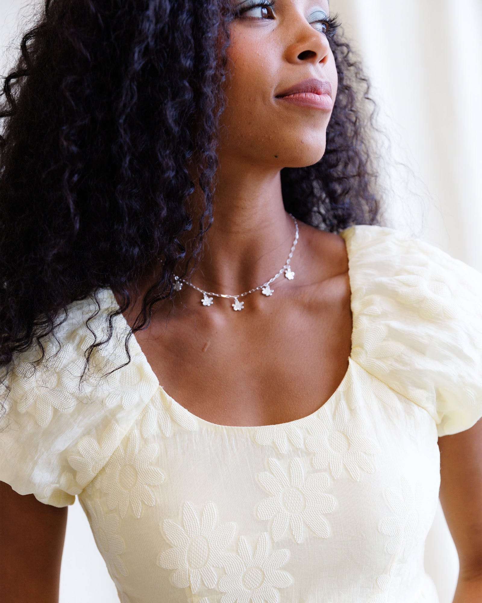 Woman wearing a white dress with puffed sleeves and a necklace, standing against a neutral background.