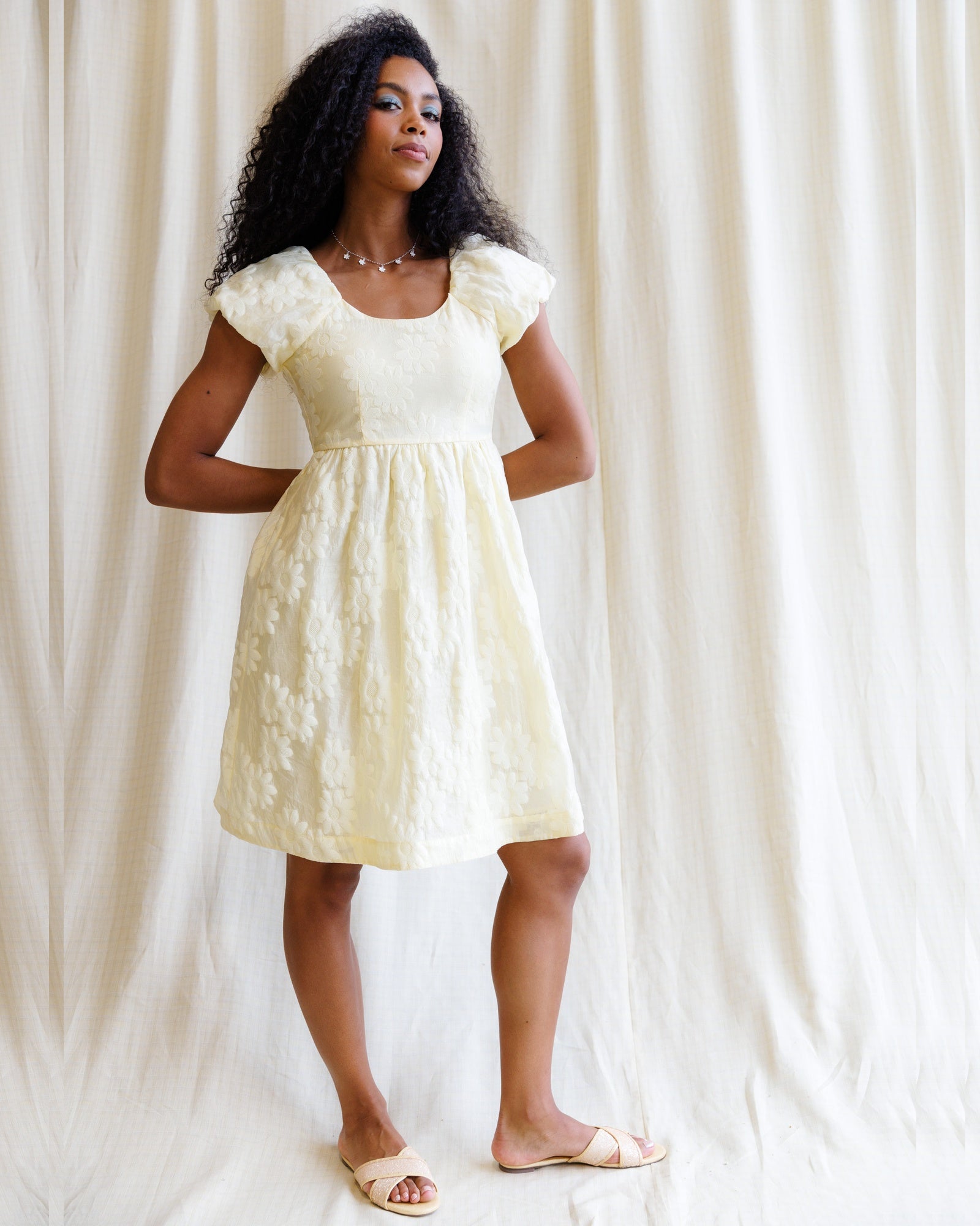 Woman wearing a light yellow dress with puffed sleeves against a white curtain background