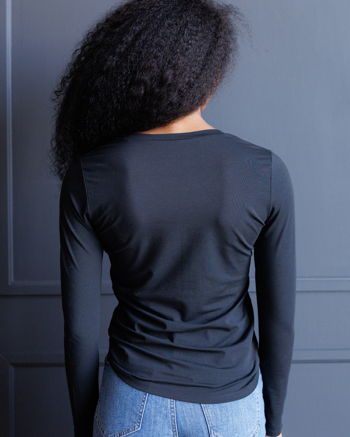 A person shown from the back wearing a fitted long-sleeve black top and light blue jeans against a dark gray paneled wall.