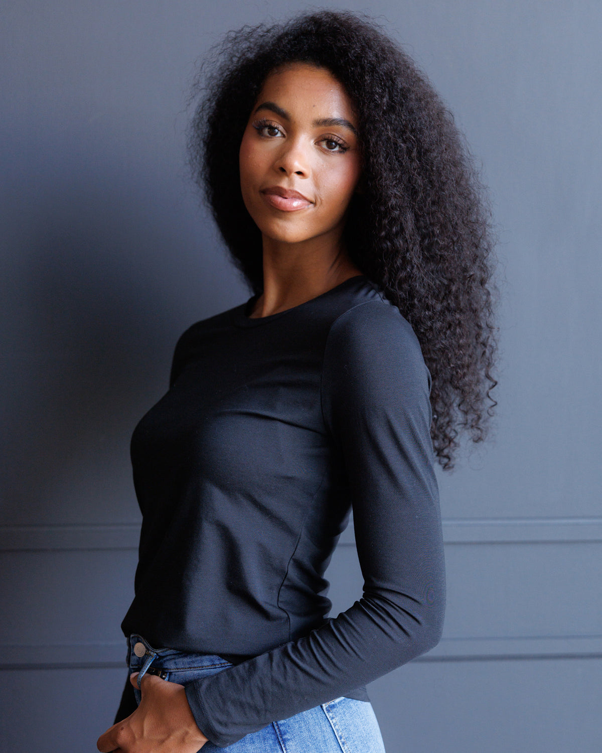 A woman with long, curly black hair wearing a fitted black long-sleeve top and blue jeans, posing against a dark gray paneled wall.