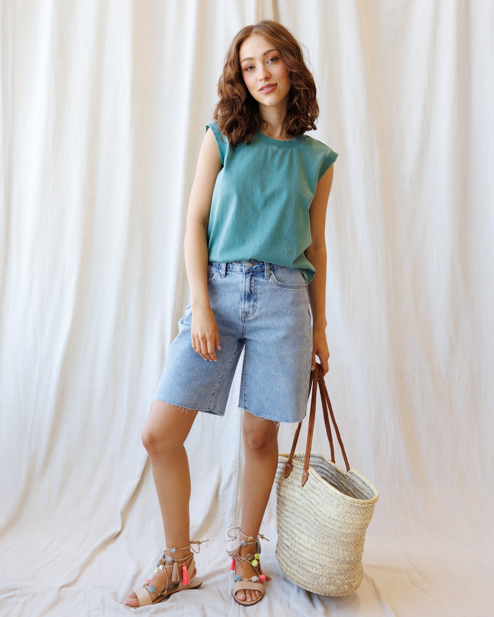 Woman wearing a teal top and denim shorts, holding a straw bag against a white curtain background