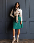 A woman stands against a dark paneled wall wearing a white long-sleeve top, a beige and cream plaid scarf, a green corduroy skirt, and beige ankle boots.