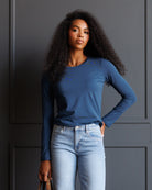 A woman with long, curly black hair wearing a slate blue long-sleeve crewneck top and light wash blue jeans, standing against a dark gray paneled wall.