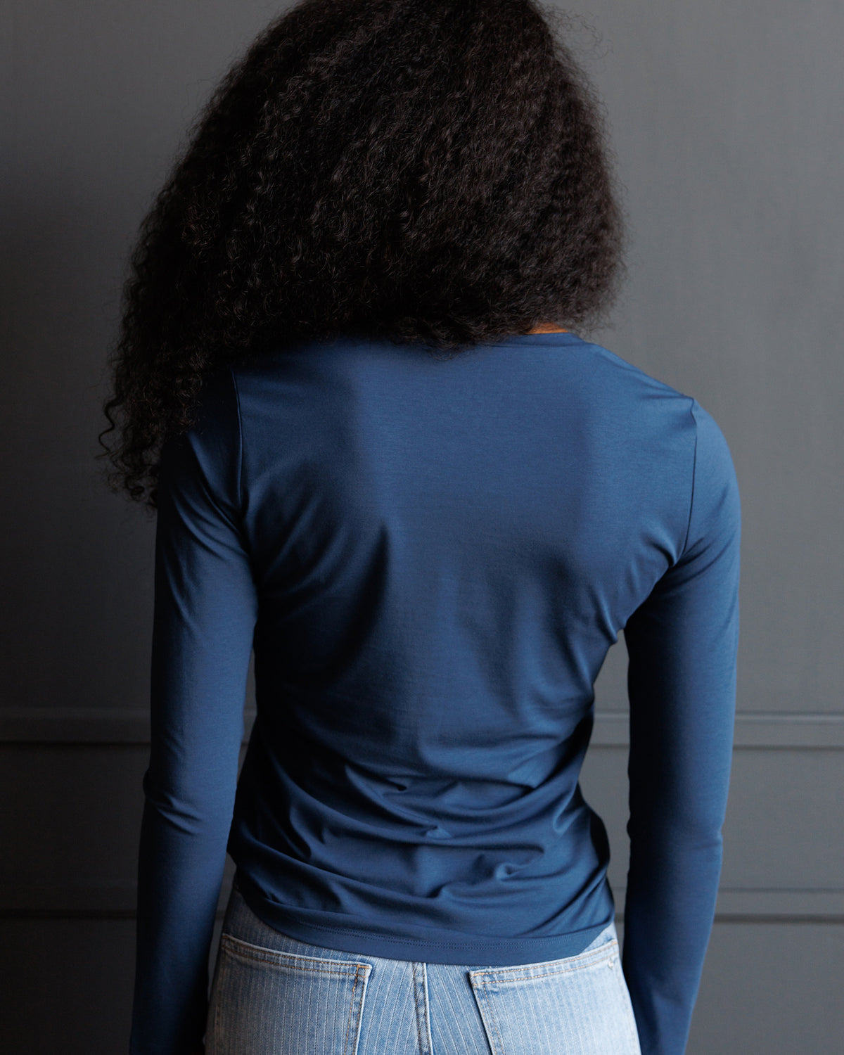 A person shown from behind wearing a fitted long-sleeve blue top and light-wash jeans against a dark gray wall.