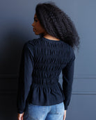 A woman with long, curly black hair is shown from the back wearing a dark navy long-sleeve top with a smocked back and light-wash blue jeans, standing against a dark blue paneled wall.