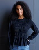 A woman with curly black hair wearing a dark navy long-sleeve smocked blouse and blue jeans, standing against a dark blue paneled wall.
