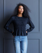 A woman with curly black hair wearing a dark navy long-sleeve smocked blouse and light blue jeans, standing against a dark blue paneled wall.