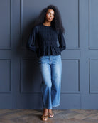 A woman with curly black hair stands against a dark blue paneled wall wearing a navy blue smocked long-sleeve top, light-wash blue jeans, and leopard-print flat sandals.