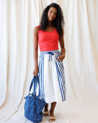 Woman wearing a red top and blue and white striped skirt holding a denim bag against a white curtain background