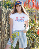 Woman wearing a white t-shirt with floral design and striped shorts, standing in a garden.
