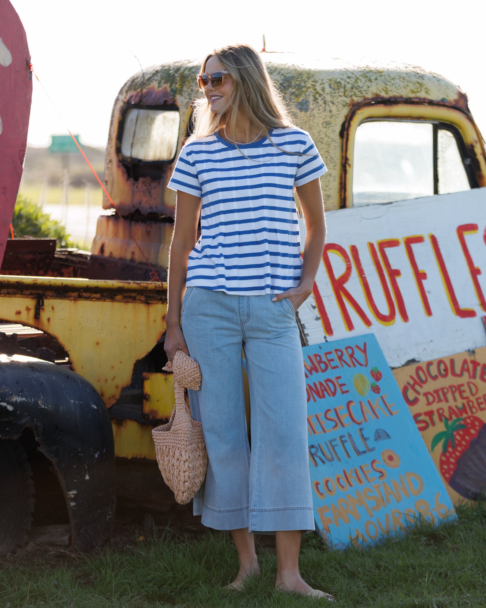 Woman in a striped shirt and light blue pants standing in front of a vintage truck and sign.