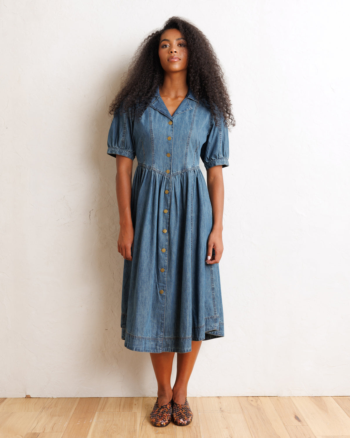 A woman modeling a short-sleeve blue denim midi dress with a button-down front, standing against a plain off-white wall on a light wood floor.