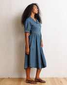 A woman wearing a short-sleeve denim midi dress with puff sleeves and a buttoned collar, standing in profile against a textured off-white wall on a light wood floor.