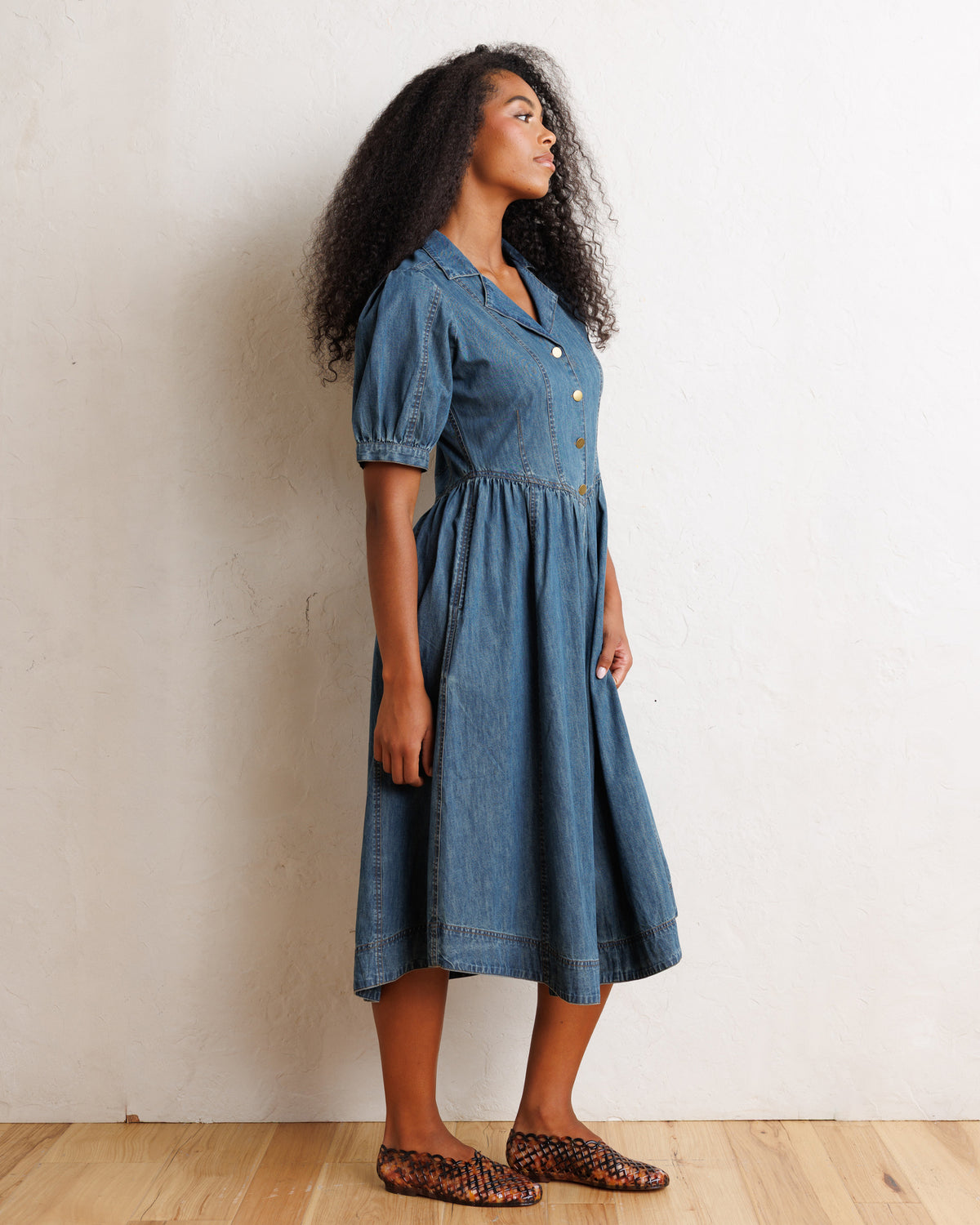 A woman wearing a short-sleeve denim midi dress with puff sleeves and a buttoned collar, standing in profile against a textured off-white wall on a light wood floor.