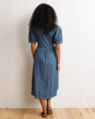 A woman with curly black hair is shown from behind wearing a blue denim midi dress with puff sleeves and a gathered waist, standing on a light wood floor against a textured off-white wall.