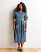 A woman modeling a short-sleeve blue denim midi dress with a button-down front, standing against a plain off-white wall on a light wood floor.