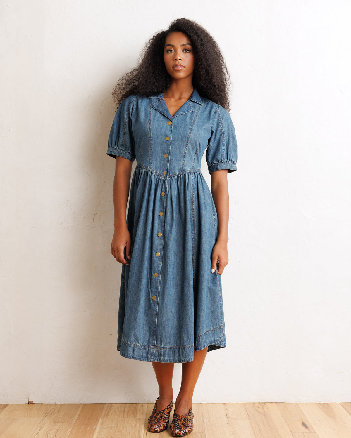 A woman modeling a short-sleeve blue denim midi dress with a button-down front, standing against a plain off-white wall on a light wood floor.