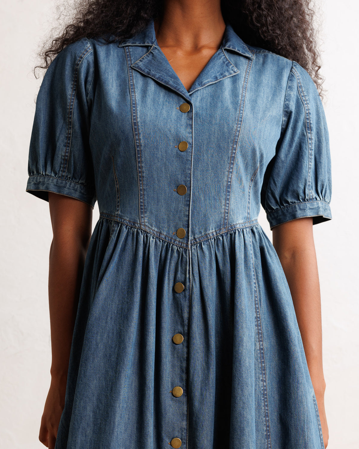 A close-up of a model wearing a blue denim shirt dress with short puff sleeves, a pointed collar, and a button-down front.
