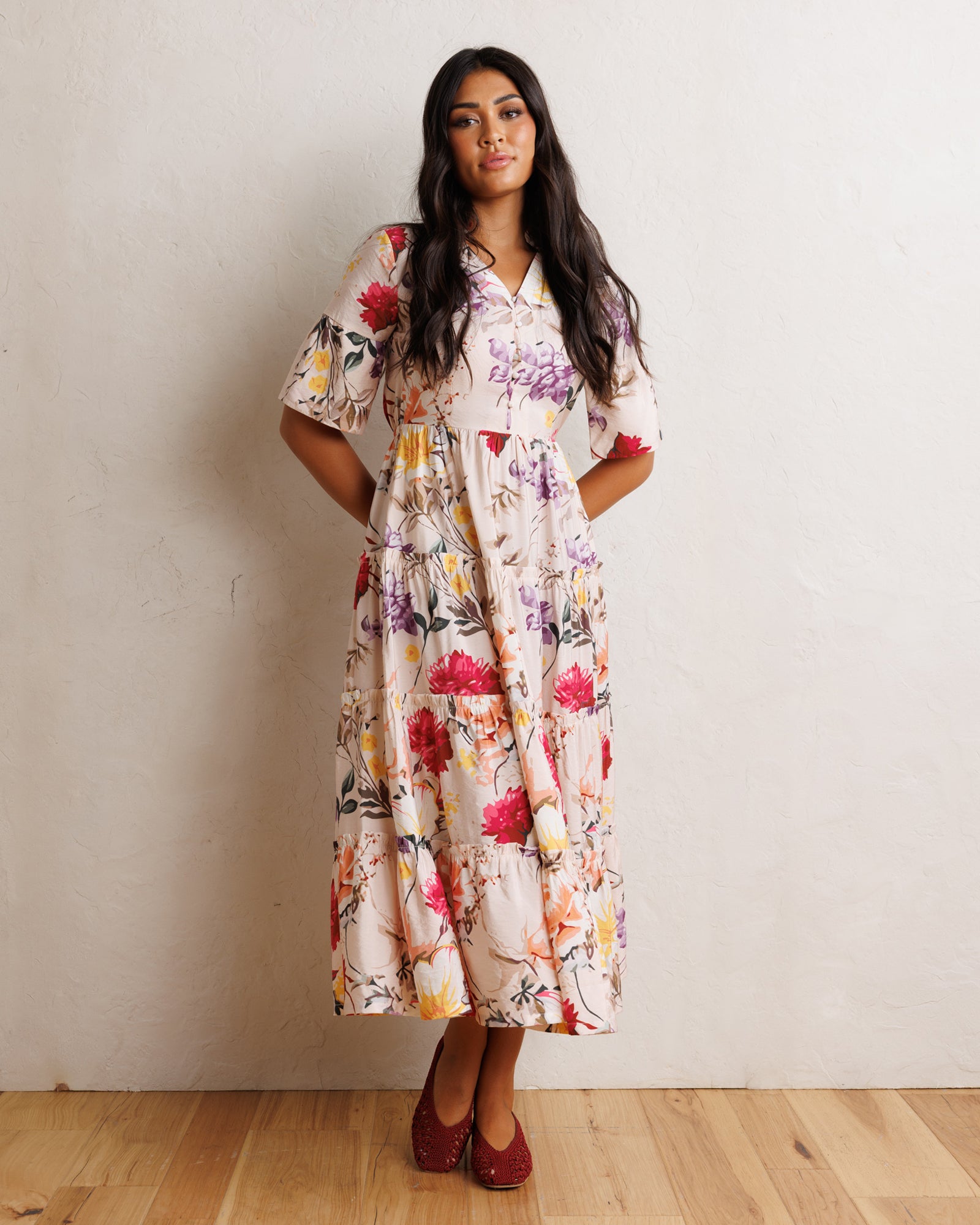 A woman modeling a short-sleeve midi dress with a colorful floral print on a cream background, standing against a textured off-white wall on a light wood floor.