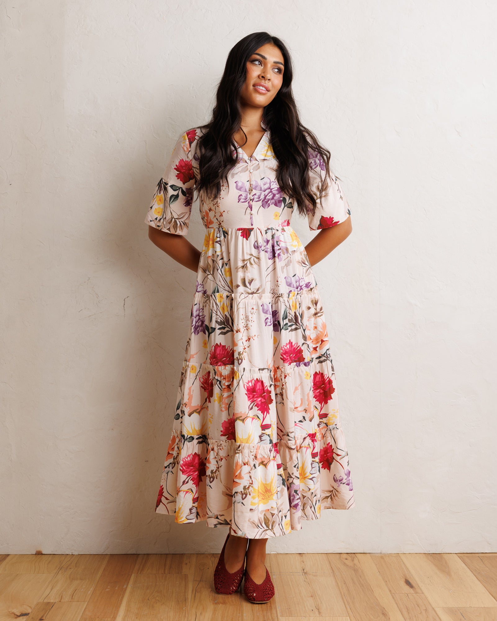A woman modeling a short-sleeve midi dress with a colorful floral print on a cream background, standing against a textured off-white wall on a light wood floor.