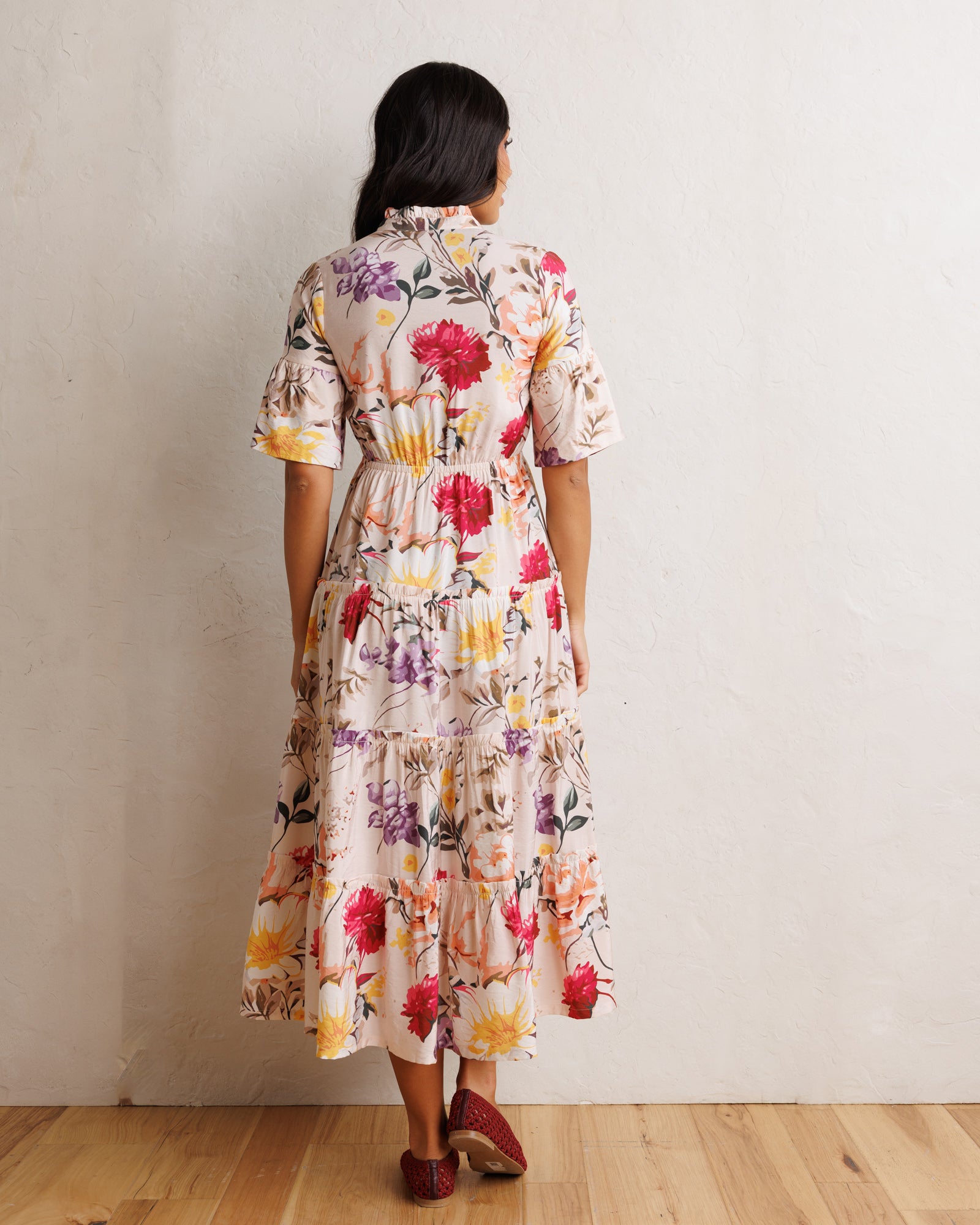 A woman viewed from behind wearing a short-sleeve, tiered midi dress with a colorful floral print on a cream background, standing against a textured off-white wall on a light wood floor.