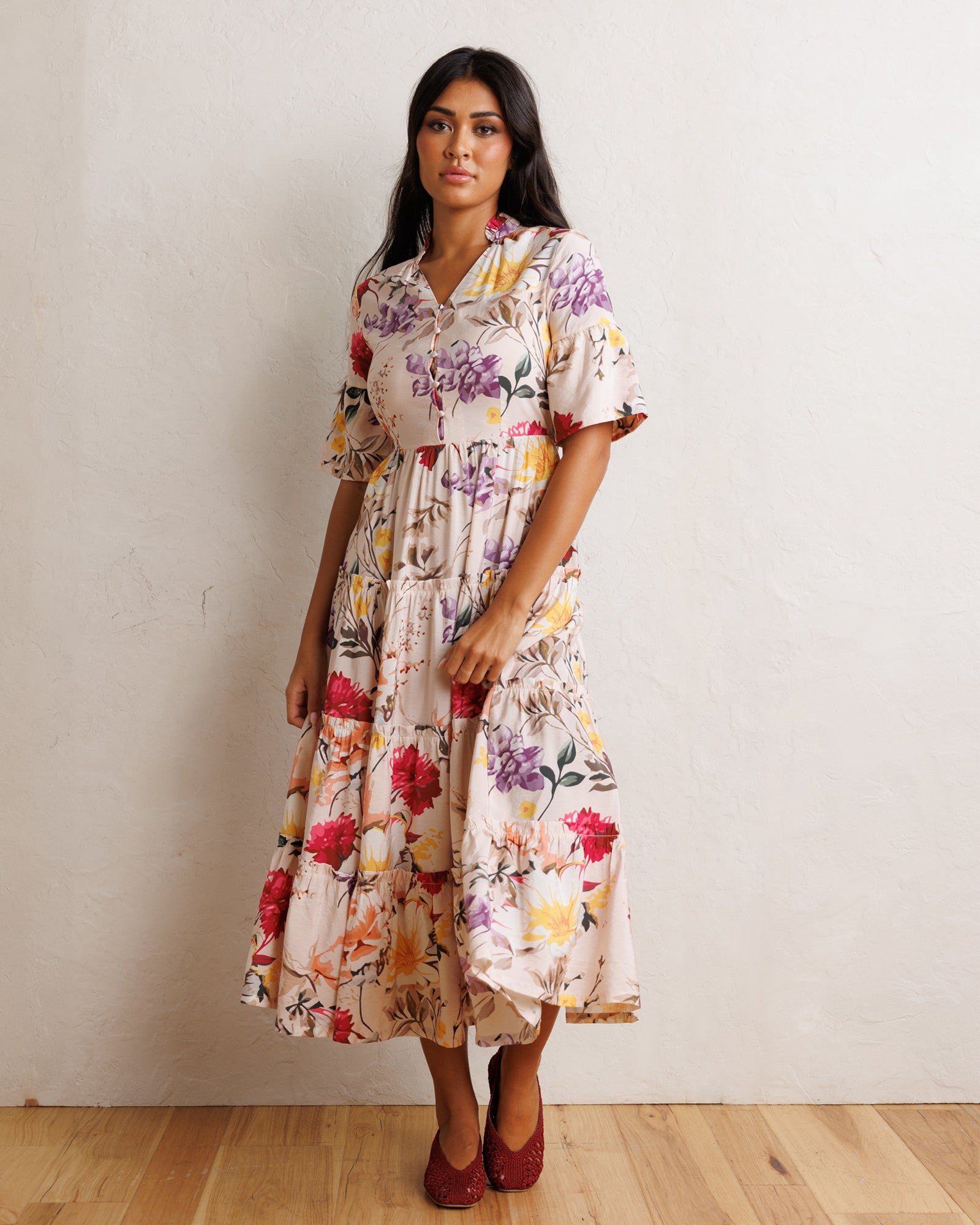 A woman models a short-sleeved midi dress with a colorful floral and botanical print on a cream background, paired with dark red pointed-toe flats, standing against a textured off-white wall and light wood floor.