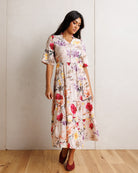 A woman modeling a short-sleeve midi dress with a colorful floral print on a cream background, standing in front of a light-colored wall and wooden floor.