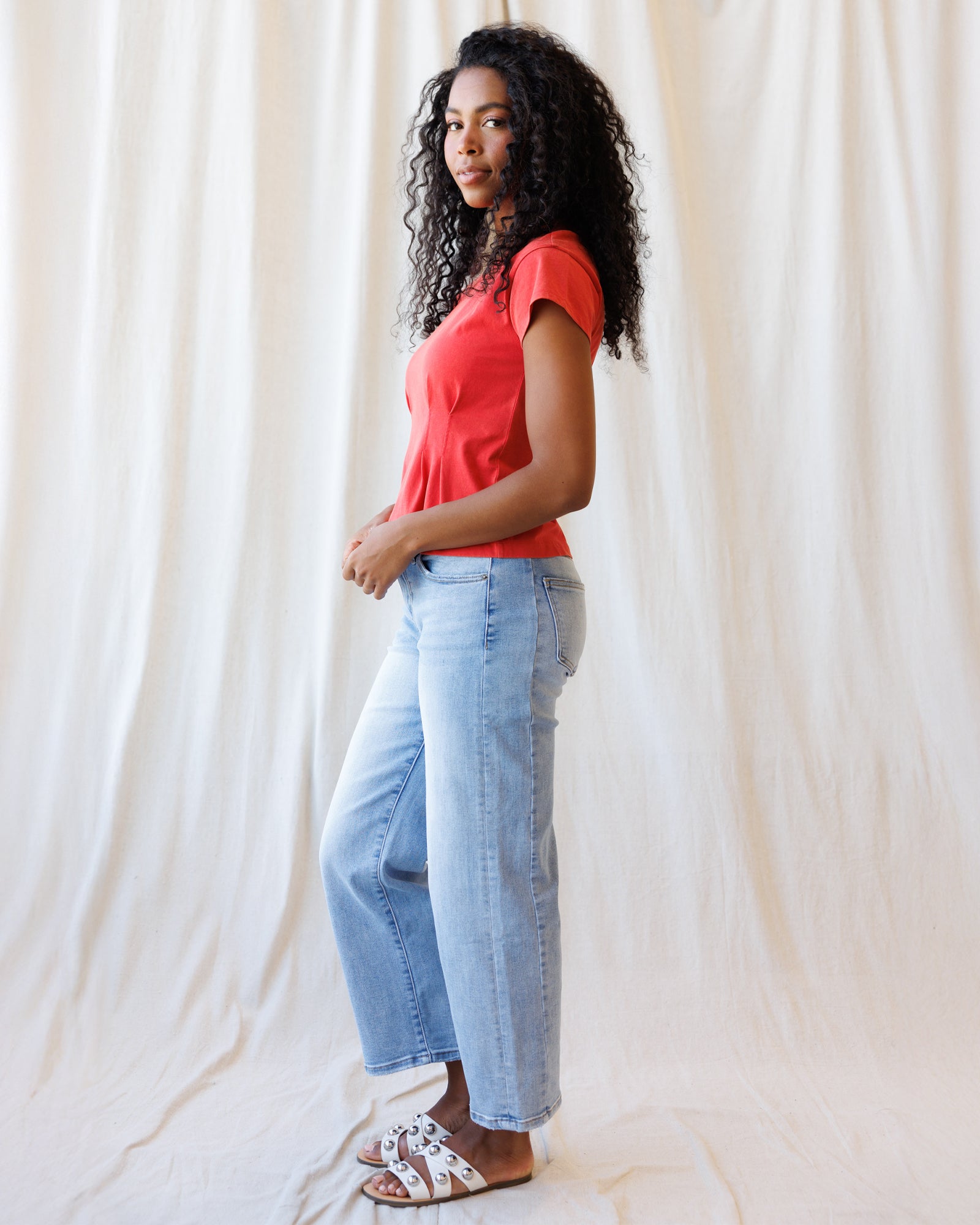 Woman wearing a red top and light blue jeans against a white curtain background