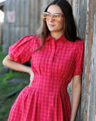 Woman in a red checkered dress and sunglasses leaning against a wooden wall.