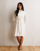 A woman modeling a white textured lace dress with long sleeves and a high collar, standing against a neutral beige wall on a light wood floor.