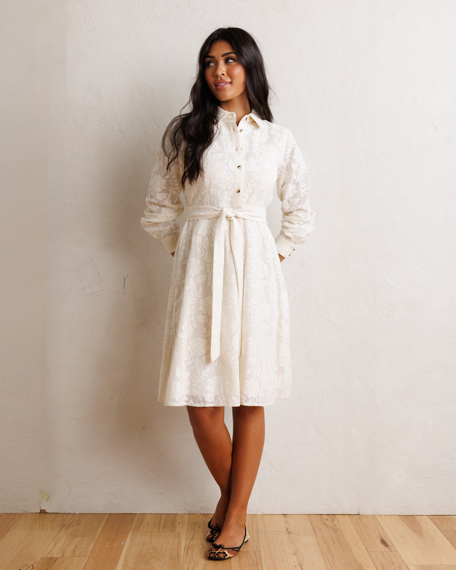 A woman modeling a white textured lace dress with long sleeves and a high collar, standing against a neutral beige wall on a light wood floor.