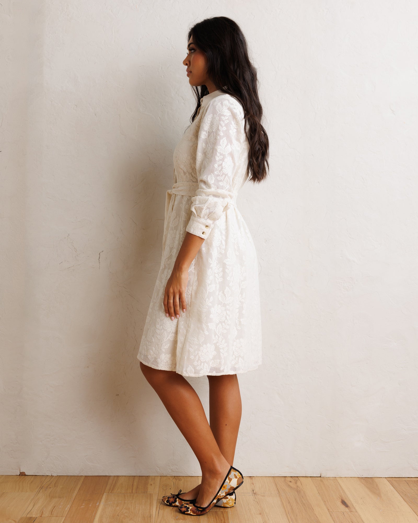 A woman wearing a white textured lace midi dress with long sleeves and a matching belt, standing in profile against a textured off-white wall on a light wood floor.