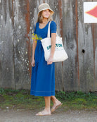 Woman in a blue dress holding a white tote bag with text, standing against a rustic wooden wall.