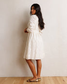 A woman wearing a white textured lace dress with long puff sleeves and a tie waist, standing against a textured off-white wall on a light wood floor.
