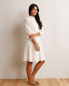 A woman modeling a white textured lace dress with long sleeves and a high collar, standing against a neutral beige wall on a light wood floor.