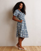 A woman modeling a short-sleeve blue and white floral midi dress with a ruffled collar, standing against a textured off-white wall on a light wood floor.