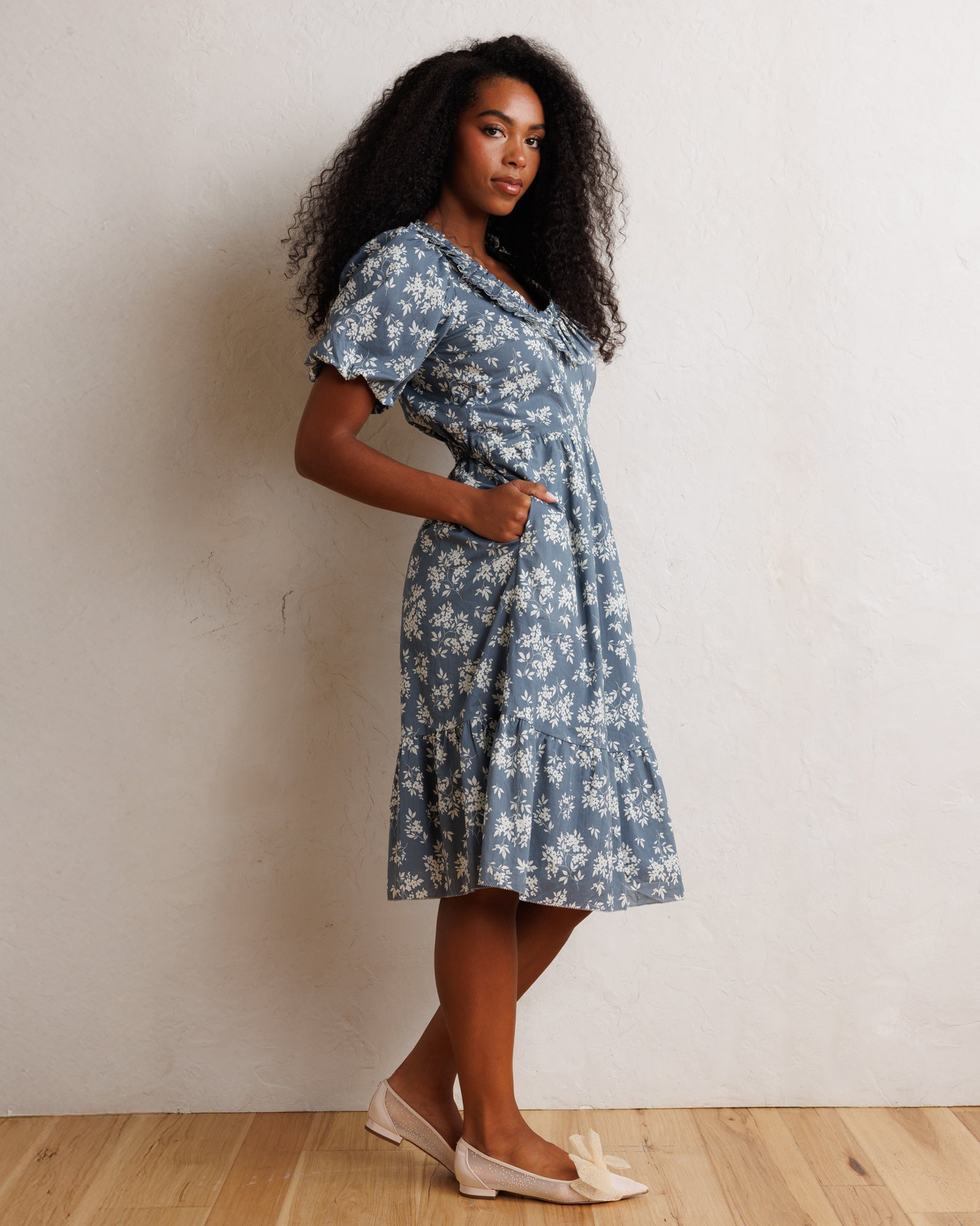 A woman modeling a short-sleeve blue and white floral midi dress with a ruffled collar, standing against a textured off-white wall on a light wood floor.
