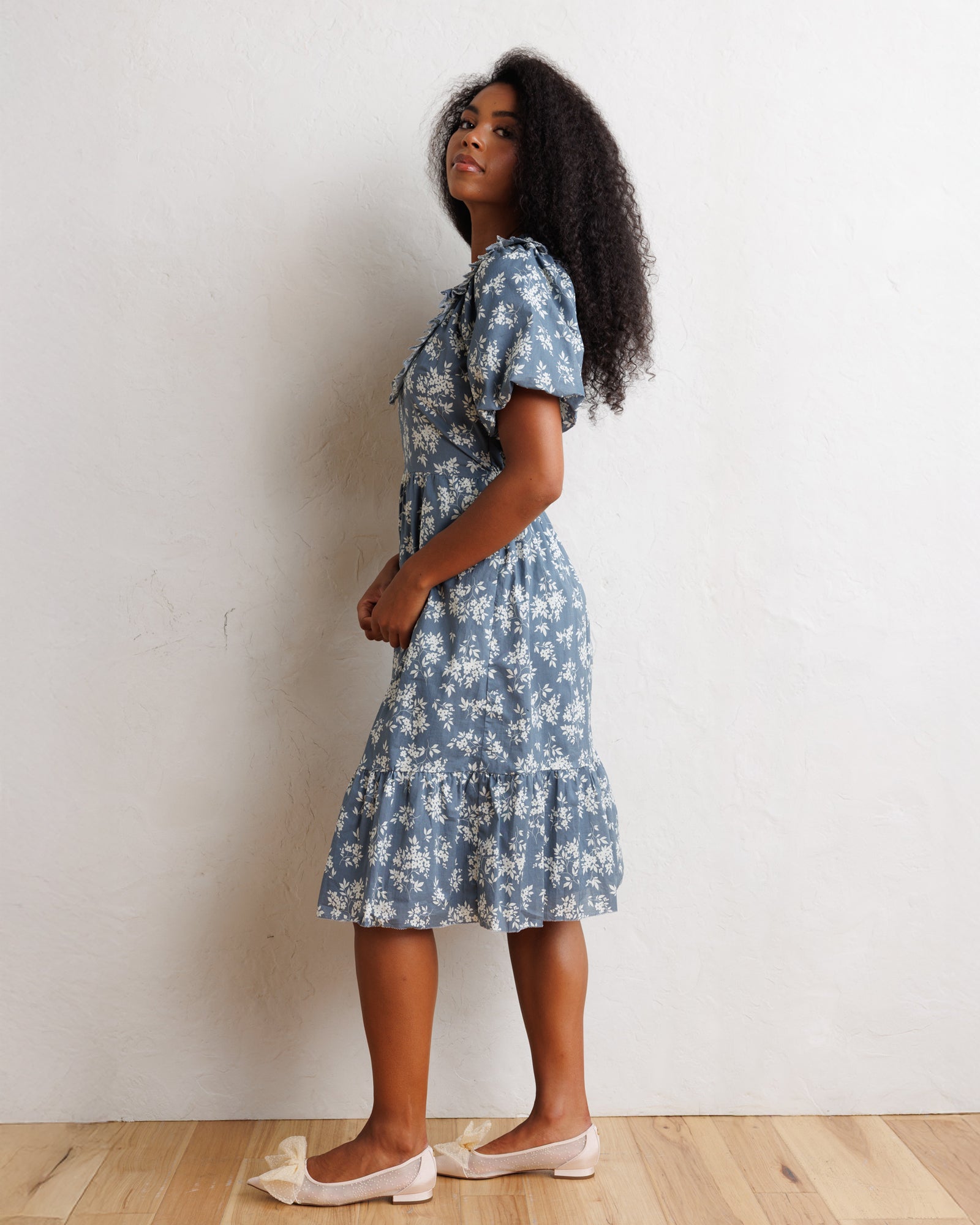 A woman stands in profile wearing a short-sleeve blue floral midi dress with a ruffled hem, paired with pale pink flats, against a plain off-white wall and light wood floor.